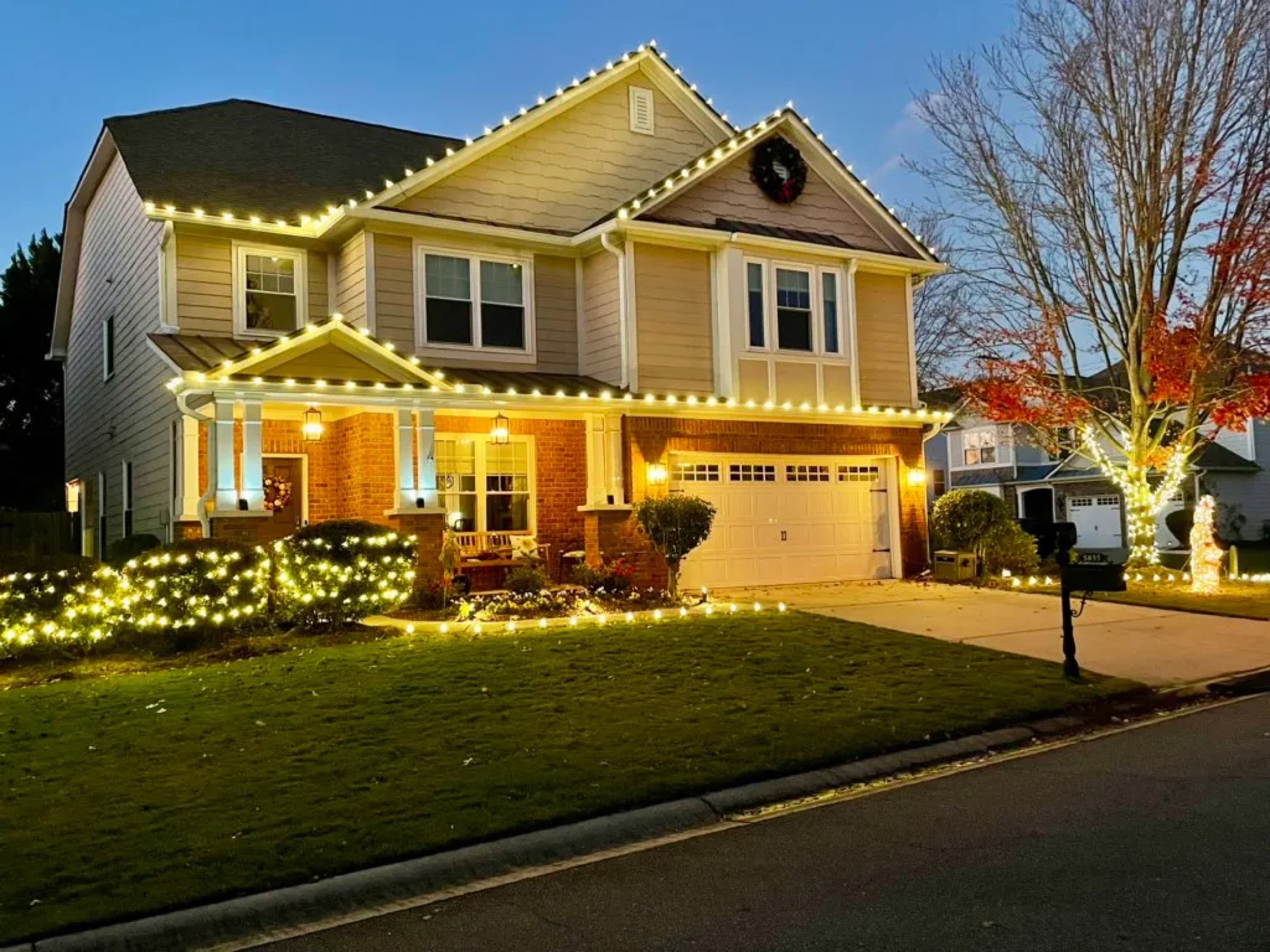 Warm white Christmas lights along the roofline, porch, and front shrubs of a two-story home at dusk